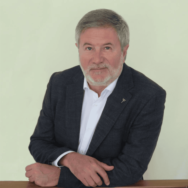 Un hombre de pelo gris y barba, vestido con traje oscuro y camisa blanca, está sentado con las manos cruzadas sobre una mesa, mirando a la cámara. Parece sereno y concentrado, probablemente preparándose para el encuentro anual de rrhh.