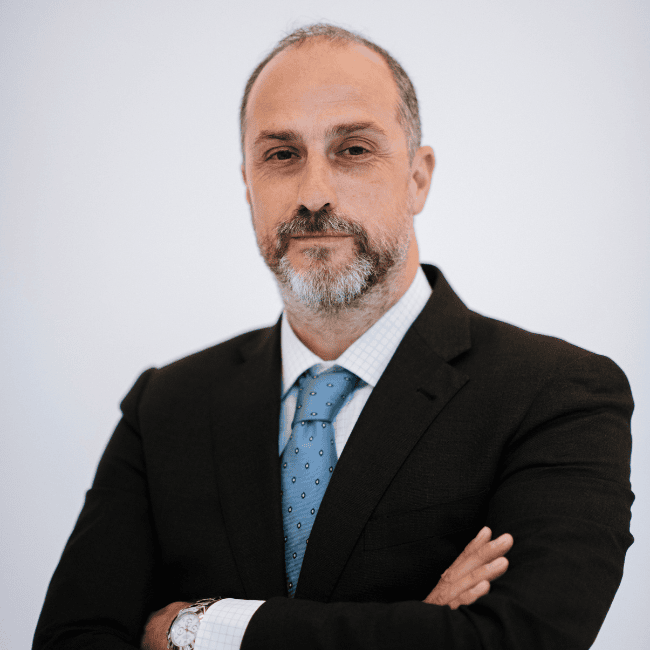Hombre de mediana edad con barba y bigote, vestido con traje y corbata azul, cruzando los brazos y mirando a la cámara sobre un fondo blanco liso, listo para el encuentro anual de rrhh.