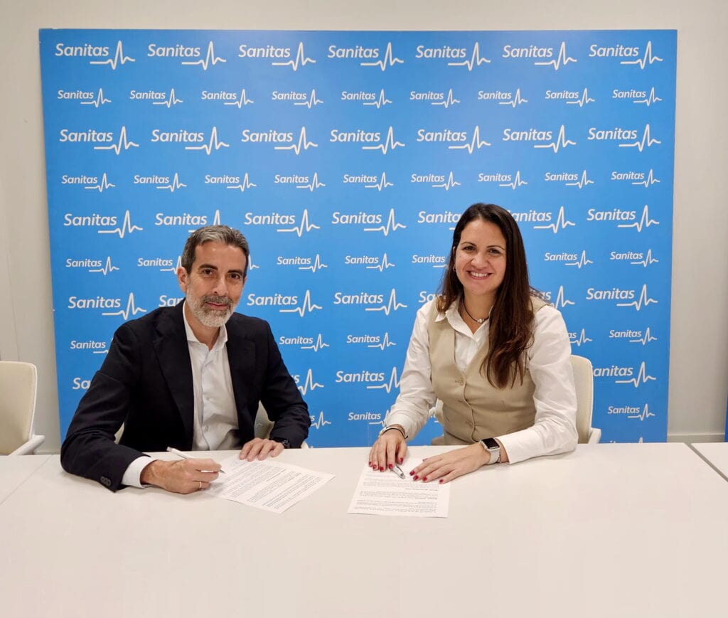 Dos personas están sentadas en una mesa con documentos frente a un fondo con la marca Sanitas, sonriendo a la cámara, mostrando su colaboración con GEHOCAN.