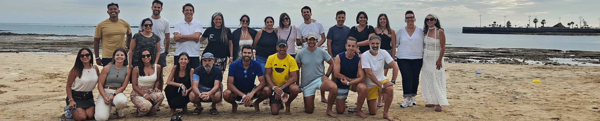 Un grupo de personas posó en una playa de arena con una costa rocosa y el océano de fondo, en un día nublado, capturando los momentos memorables de su encuentro anual de rrhh.