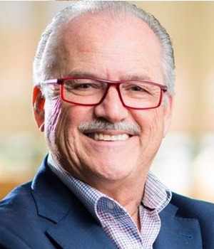 Un hombre sonriente, de pelo corto blanco y bigote, con gafas rojas, camisa a cuadros y chaqueta de traje azul oscuro; Para conocer más sobre su trabajo, contactar con gehocan.