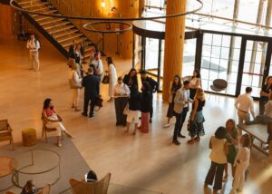 La gente se reúne y conversa en un vestíbulo moderno con techo alto, escalera y grandes ventanales. Algunos están sentados, otros de pie, en grupos.