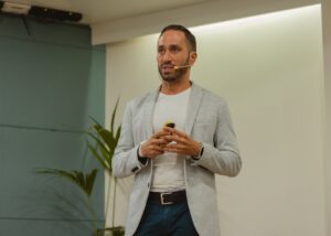 Un hombre con un blazer gris claro y una camisa blanca habla a través de un micrófono con auriculares mientras sostiene un pequeño dispositivo, de pie frente a una pared de color claro con una planta en maceta cerca.