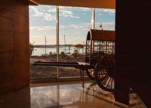 Un carro de madera se exhibe en el interior, cerca de grandes ventanales, con vista al océano y a un cielo parcialmente nublado.