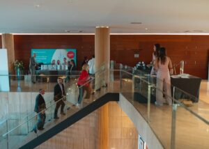 Personas caminando e interactuando en un espacio interior moderno con barandillas de vidrio y área de recepción.
