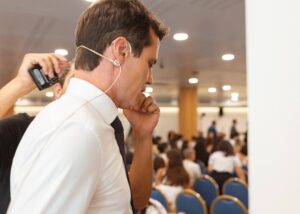 A una persona le coloca un micrófono en un auricular otro individuo en una sala de conferencias con una audiencia sentada en el fondo.
