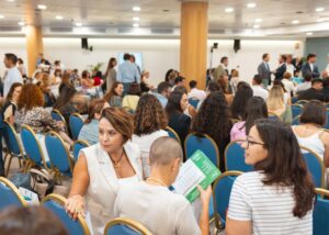 La gente está sentada en una sala de conferencias con sillas azules, conversando y sosteniendo papeles.