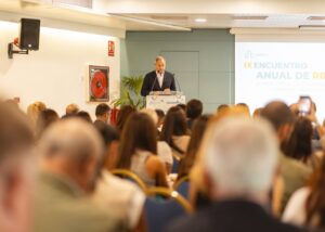 Orador en podio dirigiéndose a la audiencia durante el IX Encuentro Anual de RR, con pantalla de presentación y asistentes visibles.