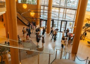 Un grupo de personas se reúne en un atrio moderno y abierto con altos pilares de madera, paredes de vidrio y lámparas circulares.