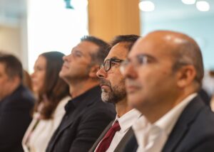 Un grupo de personas se sienta en fila y escucha atentamente. El foco está puesto en un hombre con gafas y corbata roja. El fondo está ligeramente difuminado.