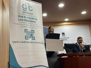 Una mujer habla en un podio junto a un hombre sentado en una sala de conferencias. Una pancarta dice "gehocan" y menciona una asociación de recursos humanos de las Islas Canarias.
