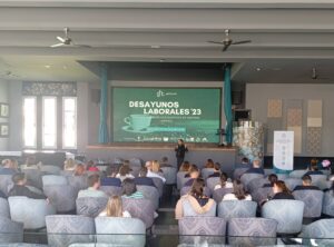 Personas sentadas en un salón frente a un escenario con una pantalla grande que muestra "Desayunos Laborales '23". Un orador se encuentra de pie al frente.