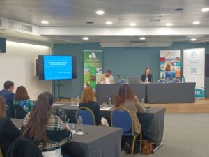 Una sala de conferencias con personas sentadas viendo una presentación en una pantalla grande. Dos oradores están sentados en una mesa al frente, con varias pancartas desplegadas en el fondo.