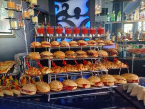 Un montaje tipo buffet con bandejas de pequeños sándwiches y bocadillos triangulares, junto a una rejilla con bebidas rojas en vasos transparentes, sobre un fondo de bar con varias botellas y decoraciones.