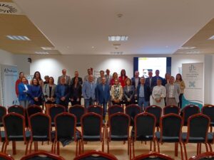 Un grupo de personas posando para una foto en una sala de conferencias con sillas vacías en primer plano. Al fondo, pancartas y una pantalla de proyección.