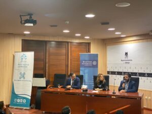 Tres personas sentadas en una mesa de conferencias con micrófonos. Al fondo se ven dos pancartas y un logotipo en la pared.