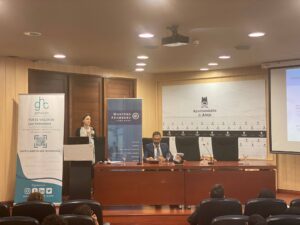 Una mujer habla en un podio junto a un hombre sentado en una sala de conferencias. Se ven diapositivas y pancartas proyectadas en el fondo.