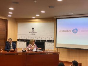 Dos personas sentadas en una mesa de conferencias en una sala de reuniones con un proyector que muestra el logotipo de Ashotel en la pantalla. Una pancarta dice "Ayuntamiento de Adeje.