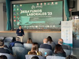 Una persona se encuentra de pie en un escenario hablando ante el público. Detrás de una gran pantalla se puede leer "Desayunos Laborales '23" sobre los cambios legislativos en materia laboral. A la derecha, una pancarta con texto.