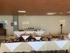 Una mesa de buffet con una variedad de pasteles, bagels y frutas, dispuesta con manteles blancos y marrones. En la parte posterior hay dispensadores de café y jugo.