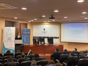 Una mujer habla en un podio en una sala de conferencias. La diapositiva de presentación dice "Claves para mejorar las cláusulas de los contratos de trabajo". Los miembros del público están sentados en filas.