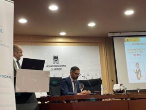 Dos hombres están sentados en una mesa de conferencias con una pantalla que muestra una presentación. Al fondo aparece un cartel que dice "Ayuntamiento de Adeje".