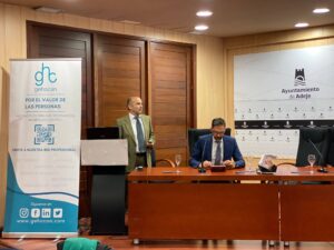 Dos hombres están en una mesa de conferencias; uno está de pie junto a una pancarta de una organización llamada Gehocan, mientras que el otro está sentado, mirando un dispositivo móvil.