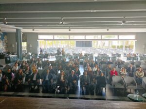 Un gran grupo de personas sentadas en filas dentro de una espaciosa sala de conferencias con grandes ventanas y ventiladores de techo.