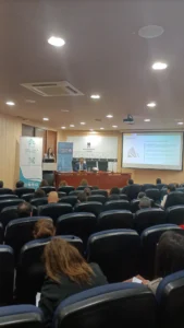 Una sala de conferencias con personas sentadas frente a un escenario, donde un orador presenta una presentación de diapositivas.