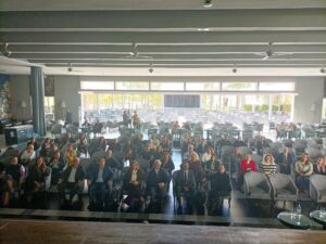 Un grupo de personas sentadas en filas de sillas frente a la cámara en una sala grande y bien iluminada con una pared de ventanas al fondo.