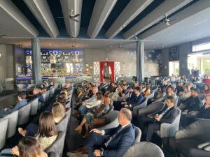 Personas sentadas en una sala espaciosa con decoración moderna, frente a un escenario o zona de presentaciones.