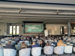 Una sala de conferencias con los asistentes sentados en filas frente a una gran pantalla que muestra "Desayunos Laborales '23". La sala cuenta con ventiladores de techo decorativos y elegantes sillones.