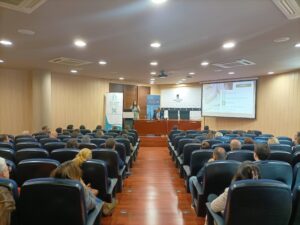 Público sentado en una sala de conferencias frente a un orador en el escenario con una pantalla de presentación.