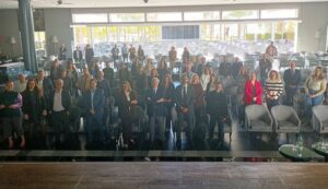 Un grupo de personas vestidas formalmente se sientan y se paran en una sala de conferencias grande y luminosa con filas de sillas y mesas.