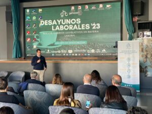 Un orador presenta en el evento "Desayunos Laborales '23". El público escucha en un ambiente tipo teatro con una gran pantalla que muestra textos y logotipos.