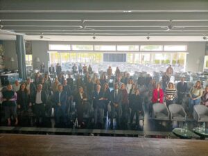 Un grupo de personas se encuentra en una sala de conferencias frente a la cámara, con filas de sillas y mesas visibles en el fondo.