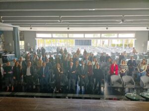 Un grupo de personas vestidas con ropa de negocios se encuentra en una sala grande y bien iluminada con muchas sillas y mesas vacías en el fondo.