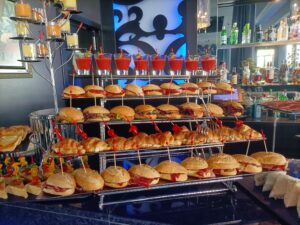 Una exhibición de varios sándwiches y bebidas dispuestos en bandejas escalonadas en un entorno de bar, con un fondo de iluminación azul y una variedad de bebidas embotelladas.