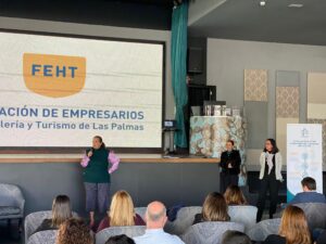 Tres personas están en el escenario en un evento empresarial con un gran proyector que muestra "FEHT" y miembros de la audiencia sentados.