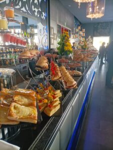 Un buffet con diversos sándwiches, brochetas y pasteles expuestos en un mostrador largo en un entorno elegante y bien iluminado.