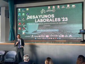 Un orador presenta en un evento titulado "Desayunos Laborales '23" con una pantalla grande que muestra información relacionada e íconos.