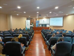 Público sentado en una sala de conferencias frente a un escenario con un orador en un podio y una presentación en una pantalla.