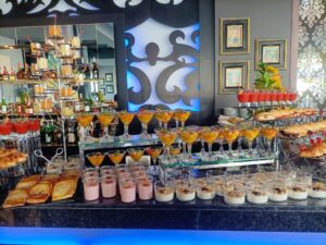 Un buffet con diversos postres y bebidas exhibidos en un stand escalonado, que incluye parfaits, pasteles y martinis cubiertos de frutas.