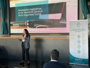 Una mujer ofrece una presentación sobre las novedades legislativas en materia de Seguridad Social, de pie junto a una pantalla grande. A la derecha se ve un cartel con información sobre el evento.