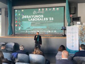 Una persona hablando en un escenario frente a una audiencia, con una gran pantalla de presentación que muestra "Desayunos Laborales '23" y varios logotipos y textos en español.
