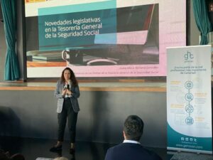 Una mujer presenta novedades legislativas en materia de seguridad social, de pie junto a una pantalla que muestra el título y un cartel que promociona a un grupo de profesionales jurídicos. Se ven miembros de la audiencia.