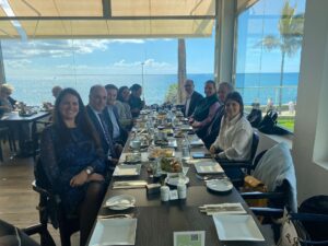 Un grupo de personas sentadas en una larga mesa de comedor, disfrutando de una comida con una brillante vista del océano de fondo.