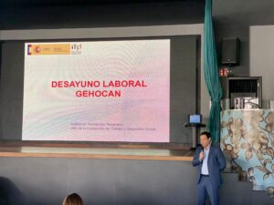 Un hombre de traje habla en el escenario con una pantalla grande que muestra una presentación titulada "Desayuno Laboral Gehocan".