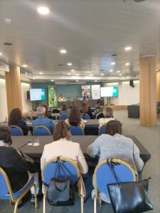 Las personas sentadas en una sala de conferencias escuchan un panel de discusión en la parte delantera. Las presentaciones se muestran en pantallas detrás de los oradores.