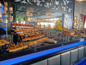 Una mesa de buffet con una variedad de sándwiches, pasteles y postres sobre un fondo de barra con varias botellas y cristalería.
